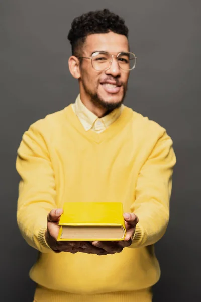 Розмитий афроамериканський чоловік в окулярах і жовтий одяг тримає книгу на темно-сірому — стокове фото
