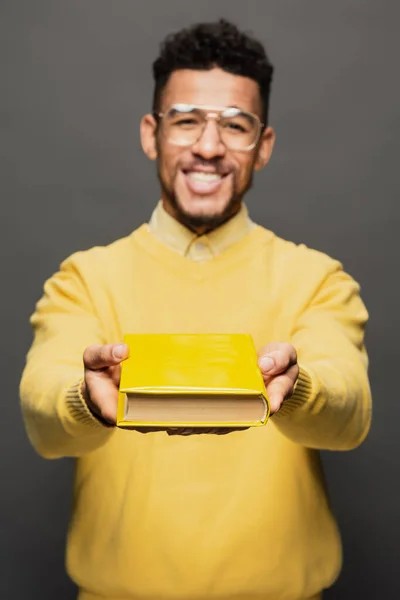 Cheerful and blurred african american man in glasses and yellow outfit holding book on dark grey — Stock Photo