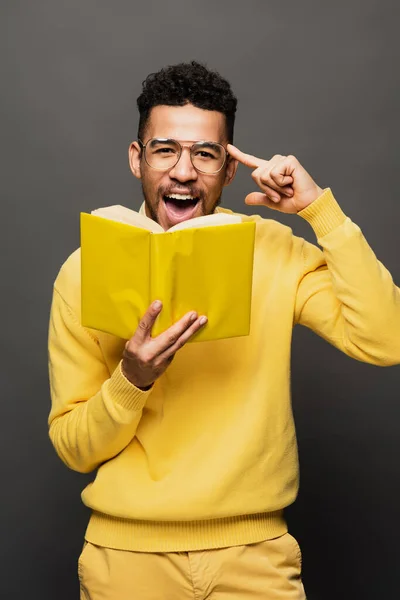 Amazed african american man in glasses and yellow outfit holding book on dark grey — Stock Photo