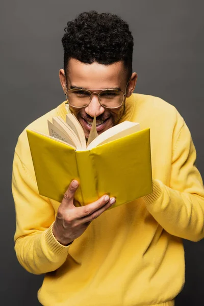 Smiling african american man in glasses and yellow outfit reading book isolated on grey — Stock Photo