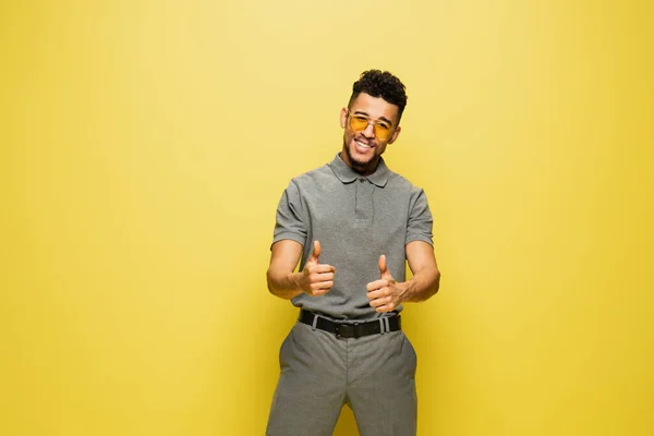 Happy african american man in sunglasses and grey tennis shirt showing thumbs up on yellow — Stock Photo