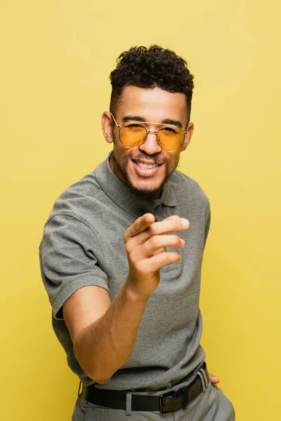 Heureux homme afro-américain en lunettes de soleil et chemise de tennis grise pointant vers la caméra isolé sur jaune — Photo de stock