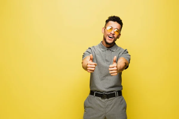 Joyful african american man in sunglasses and grey tennis shirt showing thumbs up on yellow — Stock Photo