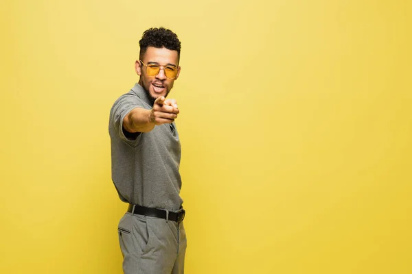 Hombre afroamericano positivo en gafas de sol y camisa de tenis gris apuntando a la cámara aislada en amarillo - foto de stock