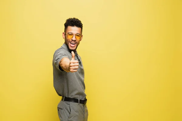Joyful african american man in sunglasses and grey tennis shirt showing thumb up on yellow — Stock Photo