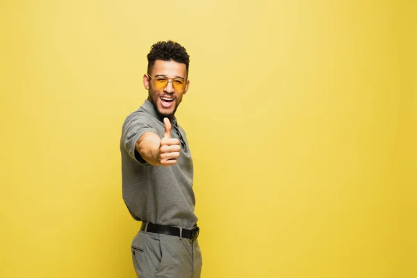 Joyeux homme afro-américain en lunettes de soleil et chemise de tennis grise montrant pouce vers le haut sur jaune — Photo de stock