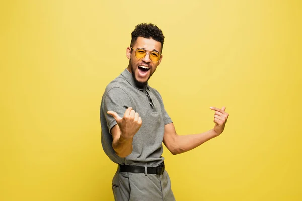Joyeux homme afro-américain en lunettes de soleil et chemise de tennis grise geste isolé sur jaune — Photo de stock
