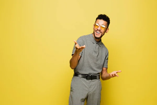 Homme afro-américain positif en lunettes de soleil et chemise de tennis grise geste isolé sur jaune — Photo de stock