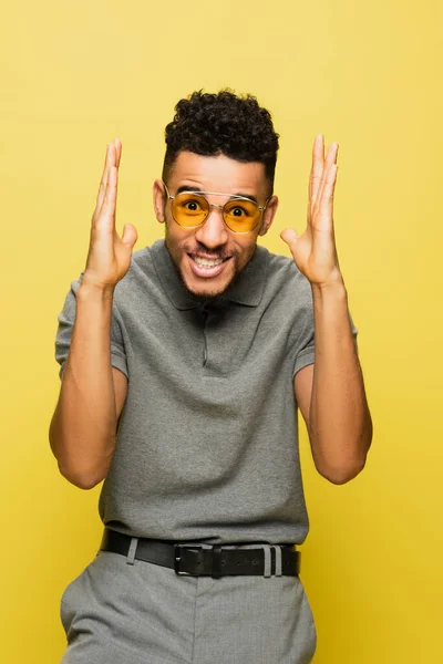 Sonriente hombre afroamericano en gafas de sol y camisa de tenis gris gesto aislado en amarillo - foto de stock