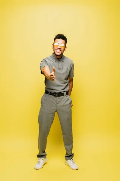 Full length of african american man in sunglasses and grey tennis shirt standing with outstretched hand on yellow — Stock Photo