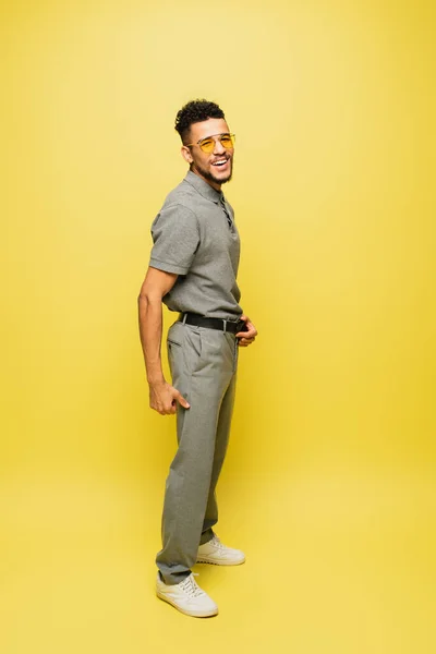 Full length of cheerful african american man in sunglasses and grey tennis shirt standing on yellow — Stock Photo