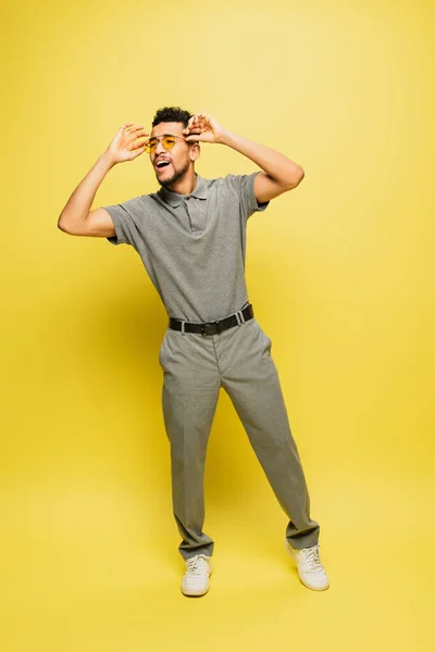 Longitud completa del hombre americano africano alegre en la camisa gris del tenis que ajusta las gafas de sol en amarillo - foto de stock