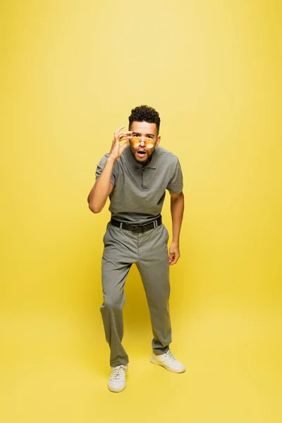 Comprimento total de homem americano africano chocado em camisa de tênis cinza ajustando óculos de sol em amarelo — Fotografia de Stock