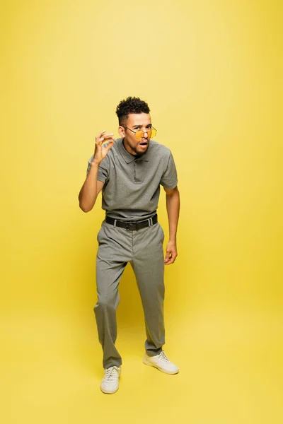 Pleine longueur de l'homme afro-américain choqué en lunettes de soleil et chemise de tennis grise debout sur jaune — Photo de stock