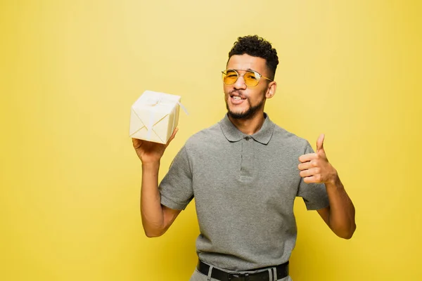 Homme afro-américain en lunettes de soleil et chemise de tennis grise tenant présent enveloppé et montrant pouce vers le haut sur jaune — Photo de stock