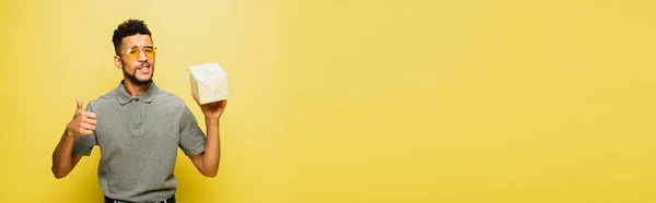 Uomo afroamericano in occhiali da sole e camicia da tennis grigia che tiene il regalo avvolto e mostra il pollice su giallo, banner — Foto stock