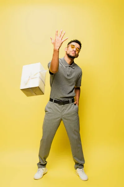 Full length of stylish african american man in sunglasses and grey tennis shirt throwing wrapped present on yellow — Stock Photo