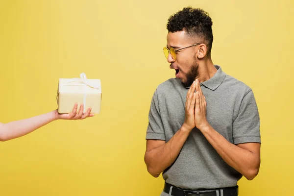 Sorprendido hombre afroamericano en gafas de sol mirando en la actualidad en mano femenina aislado en amarillo - foto de stock