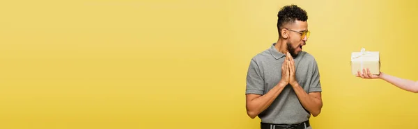 Homme afro-américain choqué dans des lunettes de soleil regardant présent dans la main féminine isolée sur jaune, bannière — Photo de stock