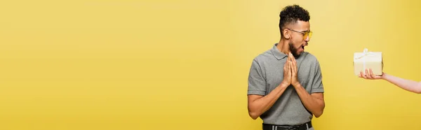 Shocked african american man in sunglasses looking at present in female hand isolated on yellow, banner — Stock Photo