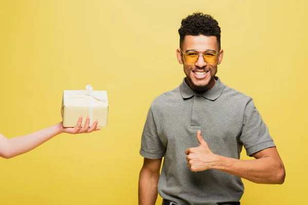 African american man in sunglasses showing thumb up near woman holding wrapped present on yellow — Stock Photo