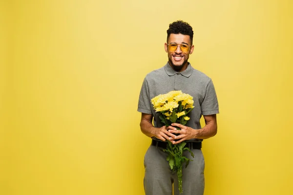 Heureux homme afro-américain en lunettes de soleil et chemise de tennis gris tenant bouquet de fleurs isolées sur jaune — Photo de stock