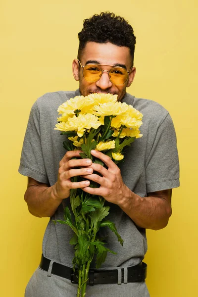 Felice uomo africano americano in occhiali da sole e camicia da tennis grigia con bouquet di fiori isolati su giallo — Foto stock