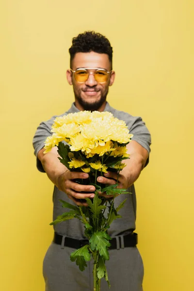 Homme afro-américain flou en lunettes de soleil et chemise de tennis grise tenant bouquet de fleurs isolé sur jaune — Photo de stock