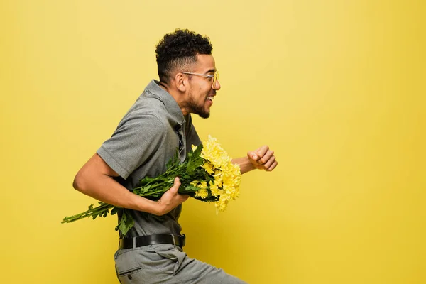 Vista lateral del hombre afroamericano feliz en gafas de sol sosteniendo ramo de flores y caminando sobre amarillo - foto de stock