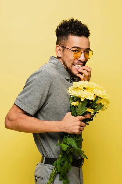 Worried african american man in sunglasses and grey tennis shirt holding bouquet of flowers isolated on yellow — Stock Photo