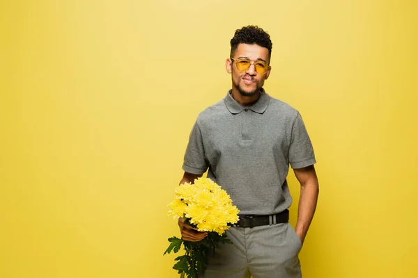 Stylish african american man in sunglasses holding bouquet of flowers and standing with hand in pocket isolated on yellow — Stock Photo