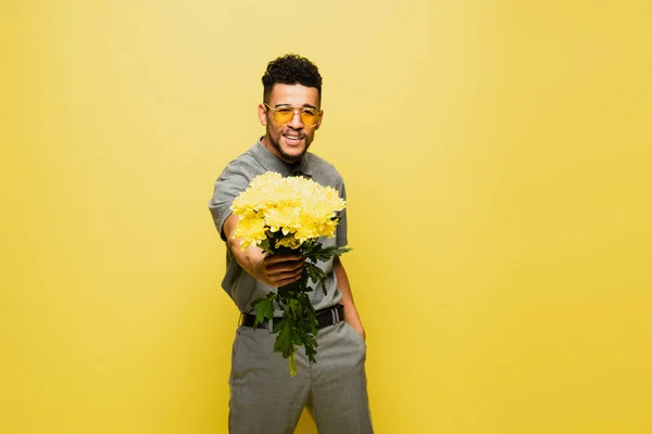 Felice uomo afroamericano in occhiali da sole che tiene bouquet di fiori e in piedi con mano in tasca isolato su giallo — Foto stock