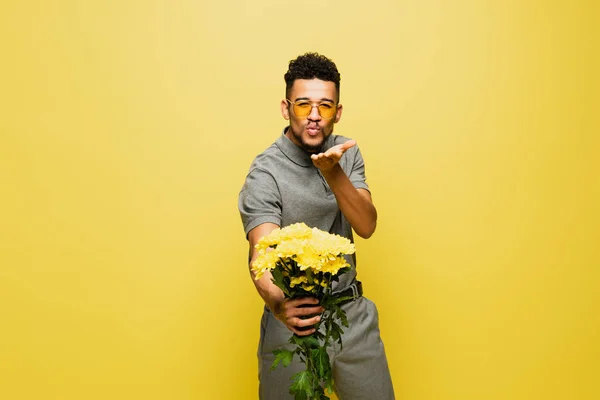 Afro-américain en lunettes de soleil tenant bouquet de fleurs et envoyant baiser d'air isolé sur jaune — Photo de stock