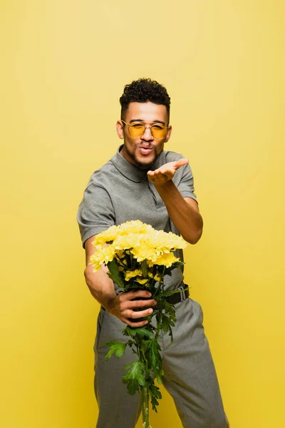 Élégant homme afro-américain en lunettes de soleil tenant bouquet de fleurs et envoyer baiser d'air isolé sur jaune — Photo de stock