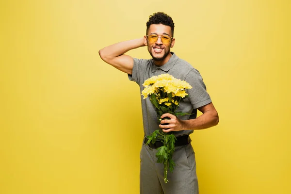 Щасливий афроамериканський чоловік в сонцезахисних окулярах тримає букет квітів на жовтому — стокове фото