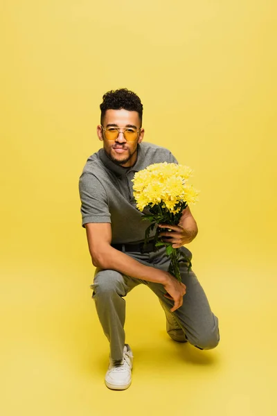 African american man in sunglasses holding bouquet of flowers and standing on knee on yellow — Stock Photo