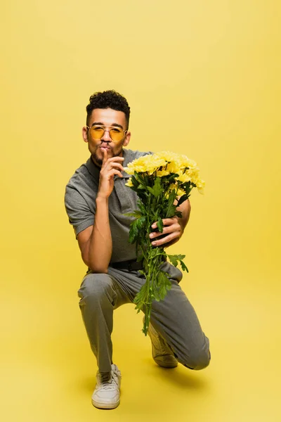 African american man in sunglasses holding bouquet of flowers while standing on knee and showing hush sign on yellow — Stock Photo