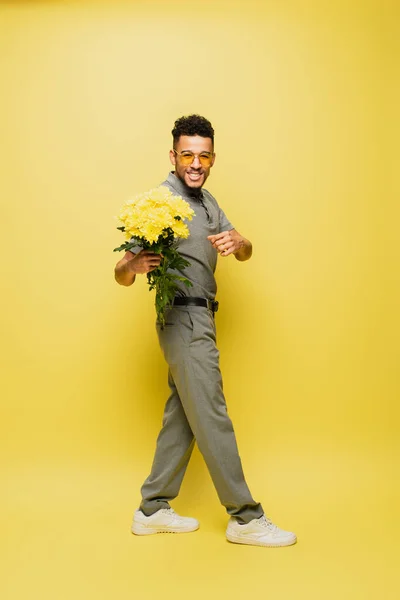 Longitud completa de hombre afroamericano feliz en gafas de sol con ramo de flores y de pie en amarillo - foto de stock