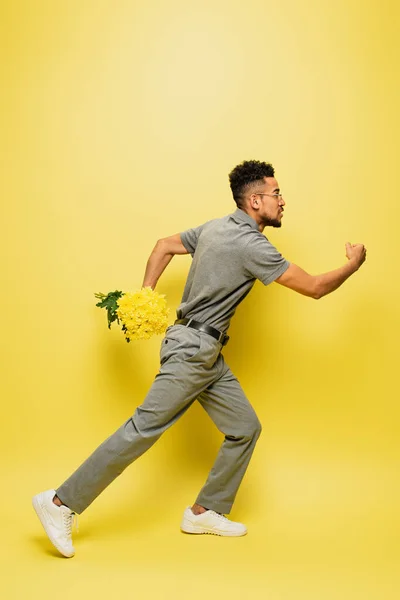 Vue latérale de l'homme afro-américain en lunettes de soleil tenant bouquet de fleurs et courant sur jaune — Photo de stock