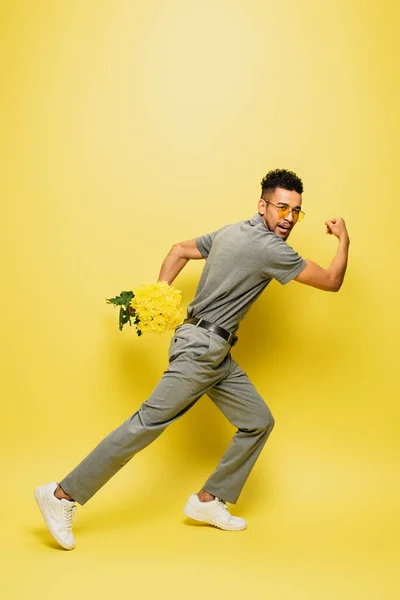 Full length of african american man in sunglasses holding bouquet of flowers and running on yellow — Stock Photo