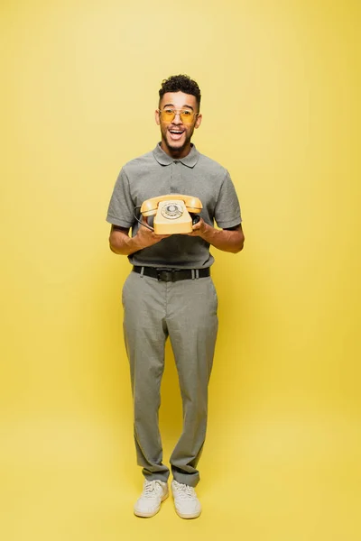 Longitud completa de hombre afroamericano excitado en gafas de sol y camisa de tenis gris sosteniendo teléfono retro en amarillo - foto de stock