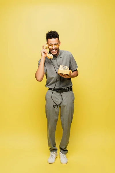 In voller Länge glücklicher afrikanisch-amerikanischer Mann in Sonnenbrille und grauem Tennishemd, der am Retro-Telefon auf gelb spricht — Stockfoto