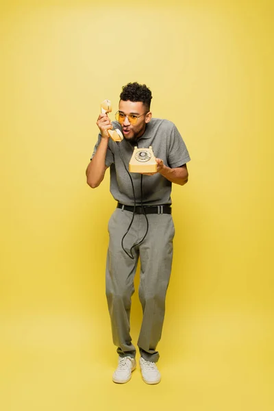 Pleine longueur de l'homme afro-américain choqué en lunettes de soleil et chemise de tennis grise parler sur téléphone rétro sur jaune — Photo de stock