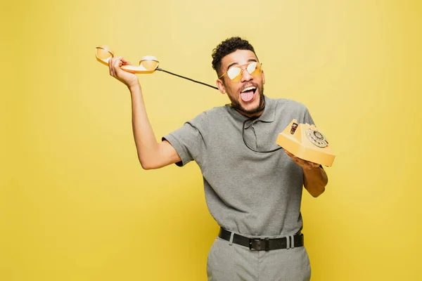 Homem americano africano em óculos de sol saindo da língua enquanto se engasga com cabo de telefone em amarelo — Fotografia de Stock