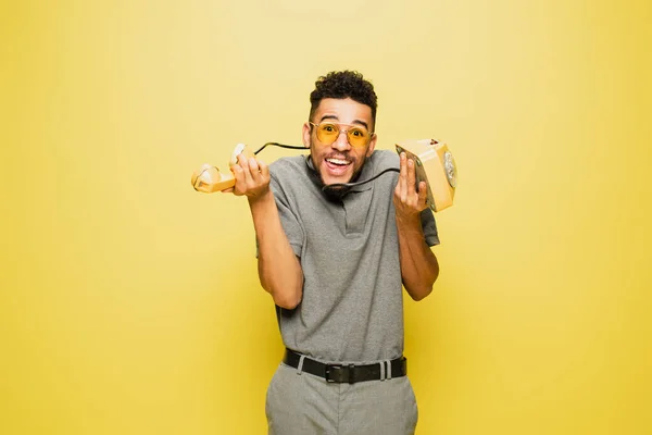 Confused african american man in sunglasses and grey tennis shirt holding retro telephone on yellow — Stock Photo