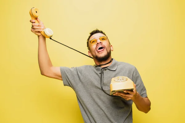 Jovem afro-americano em óculos de sol saindo da língua enquanto se engasga com cabo de telefone em amarelo — Fotografia de Stock