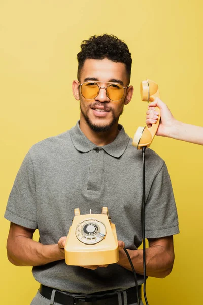 Mujer sosteniendo el teléfono cerca de hombre afroamericano en gafas de sol y camisa de tenis gris aislado en amarillo - foto de stock