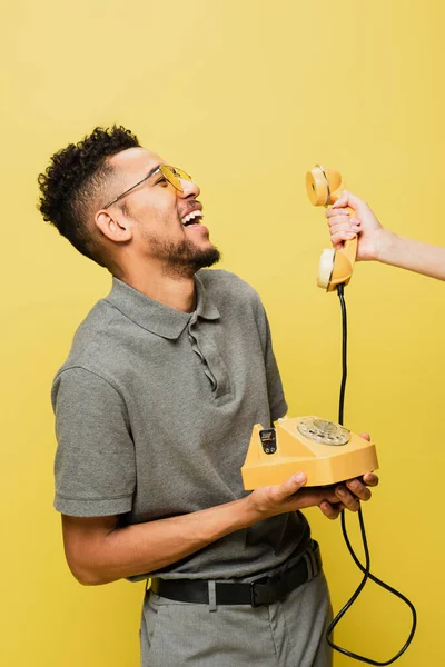 Mujer sosteniendo el teléfono cerca del hombre afroamericano positivo en gafas de sol y camisa de tenis gris aislado en amarillo - foto de stock