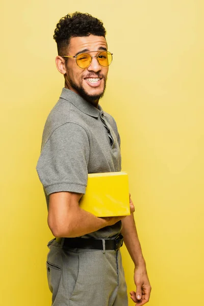 Glücklicher afrikanisch-amerikanischer Mann mit Sonnenbrille und grauem Tennishemd, Buch isoliert auf gelb — Stockfoto
