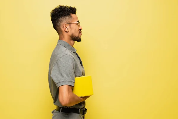 Vue latérale de heureux homme afro-américain en lunettes de soleil et chemise de tennis gris tenant livre isolé sur jaune — Photo de stock